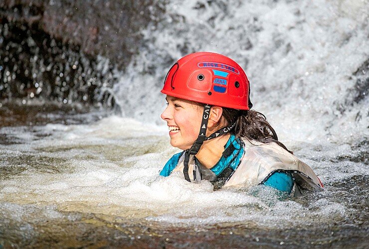 A pupil wearing a red helmet smiles while swimming in a river during an outdoor adventure activity.