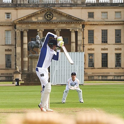 A batter swinging his bat on a cricket pitch with a wicketkeeper in position outside the school