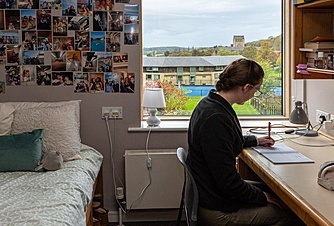 Girl sitting at a table in the process of writing with bed in the background