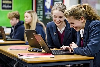Two girls in school uniform looking at a laptop schreen, smiling
