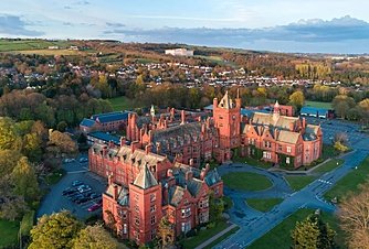 Aerial view of a large red-brick school complex set among trees with a town and rolling hills in the background.