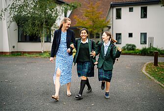 An adult woman walks with two pupils in school uniform along a paved path on campus.