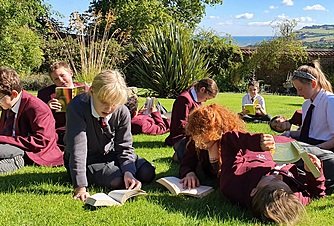 Students in school uniforms reading books while sitting and lying on the grass in the sunshine.