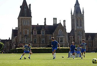 Pupils practising football on a green field with an impressive historic school building in the background.