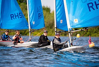 Pupils sailing small boats with bright blue sails on a calm lake, concentrating as they steer and adjust their ropes.