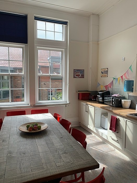 A kitchen with white cupboards, a microwave, a kettle and a dining table with red chairs.
