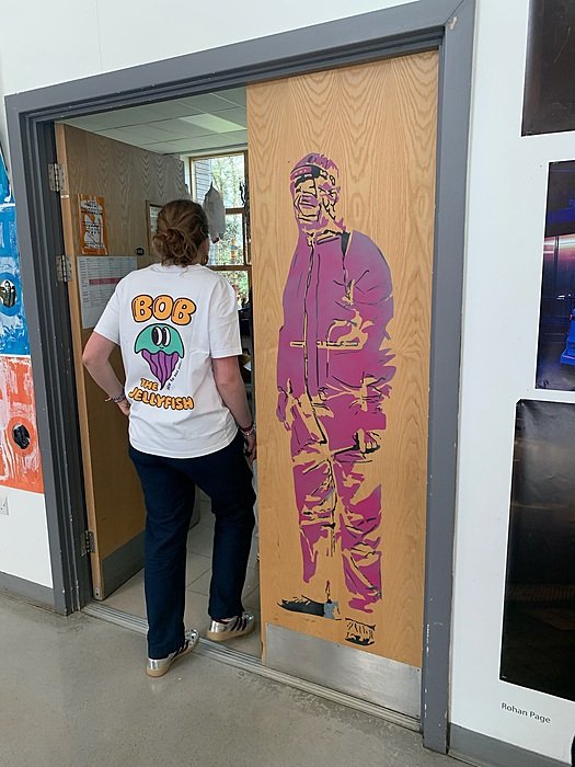 A person in a white T-shirt stands by a doorway next to a pink and yellow figure painted on the wall.
