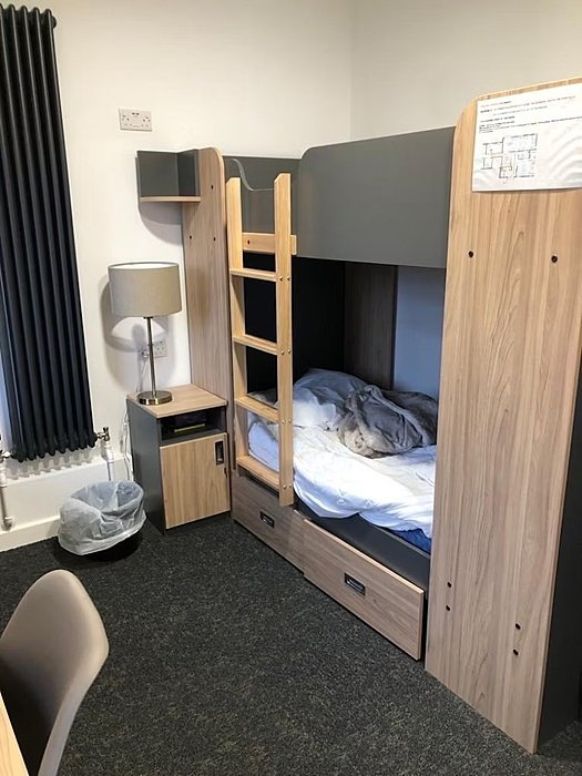 Student bedroom with wooden bunk beds, a lamp, and a small bedside table.