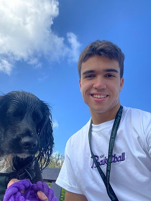 A young man takes a selfie outdoors with a black dog on a bench under a bright blue sky.
