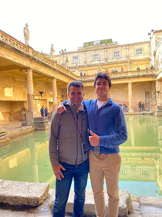 Two people stand arm in arm at a historic bath site with stone architecture and green water.