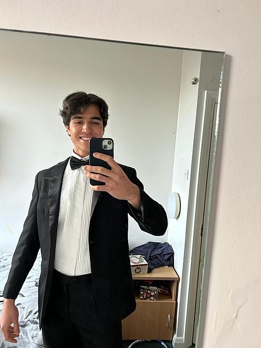 A young man takes a mirror selfie in his room, dressed in a black tuxedo with a bow tie and white shirt.