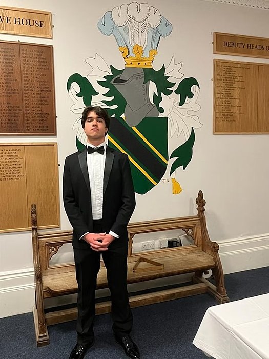 A student dressed in a black tuxedo stands in a school hall with a large coat of arms on the wall behind him.