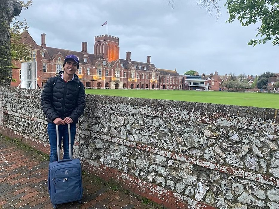 A student stands with luggage in front of a large red-brick school building with a tower and green field.