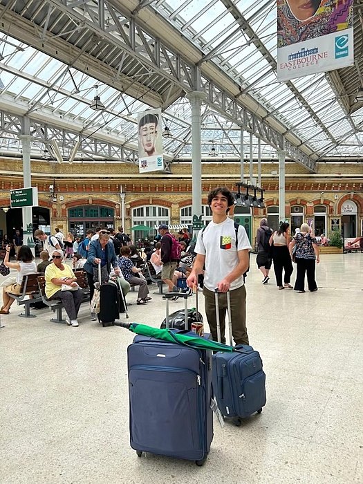A young person stands in a busy train station with a large glass roof, holding two blue suitcases.