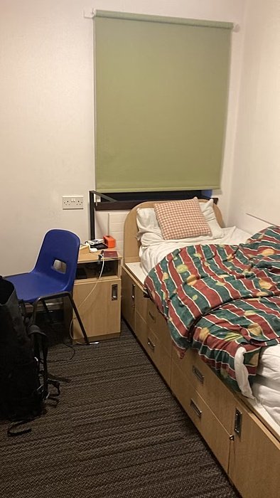 A small dorm room with a single bed, green blinds, a blue chair, and a wooden desk beside the bed.