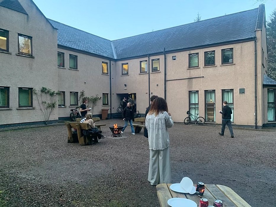 Students gather in a courtyard between dormitory buildings, sitting around a small fire pit and chatting in the evening light.