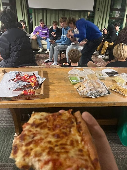 A group of students sit together indoors enjoying food, with pizza, snacks, and drinks on a wooden table.