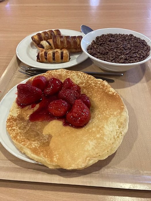 A breakfast tray with a pancake topped with strawberries, a bowl of chocolate cereal, and pastries on plates.