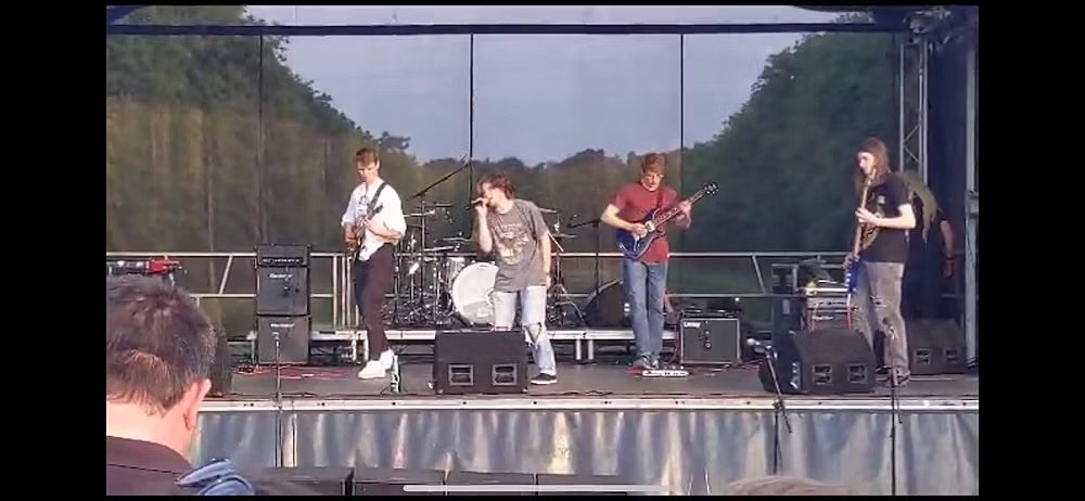 A band performs on an outdoor stage, with musicians playing guitars, drums, and singing in front of trees.