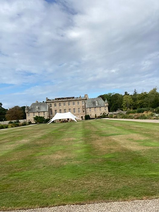 A grand stone manor house with a large lawn and a white canopy tent set up in front under a cloudy sky.