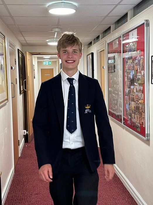 Student in school uniform standing in a brightly lit corridor lined with framed pictures and noticeboards.