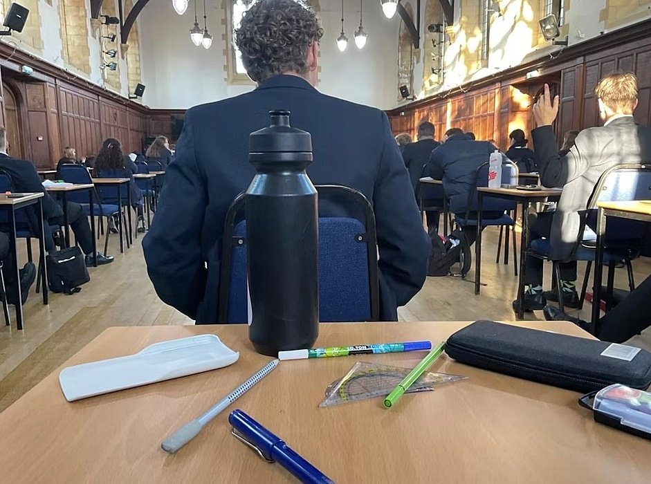 View from a desk inside an exam hall with students seated at tables and a water bottle in the foreground.