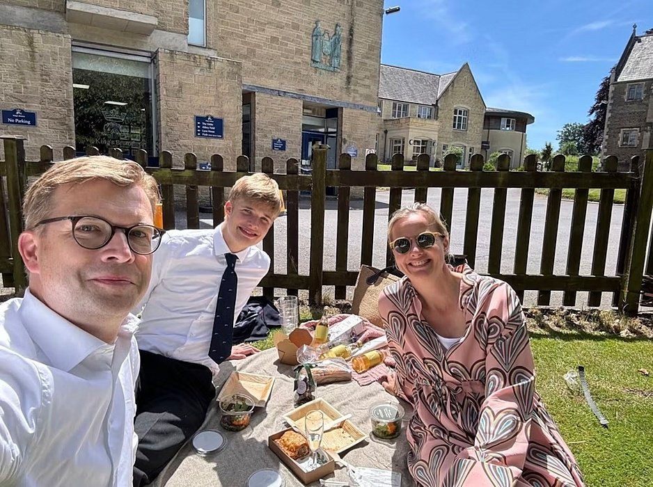 Three people enjoying a picnic on the grass outside a school building on a sunny day.