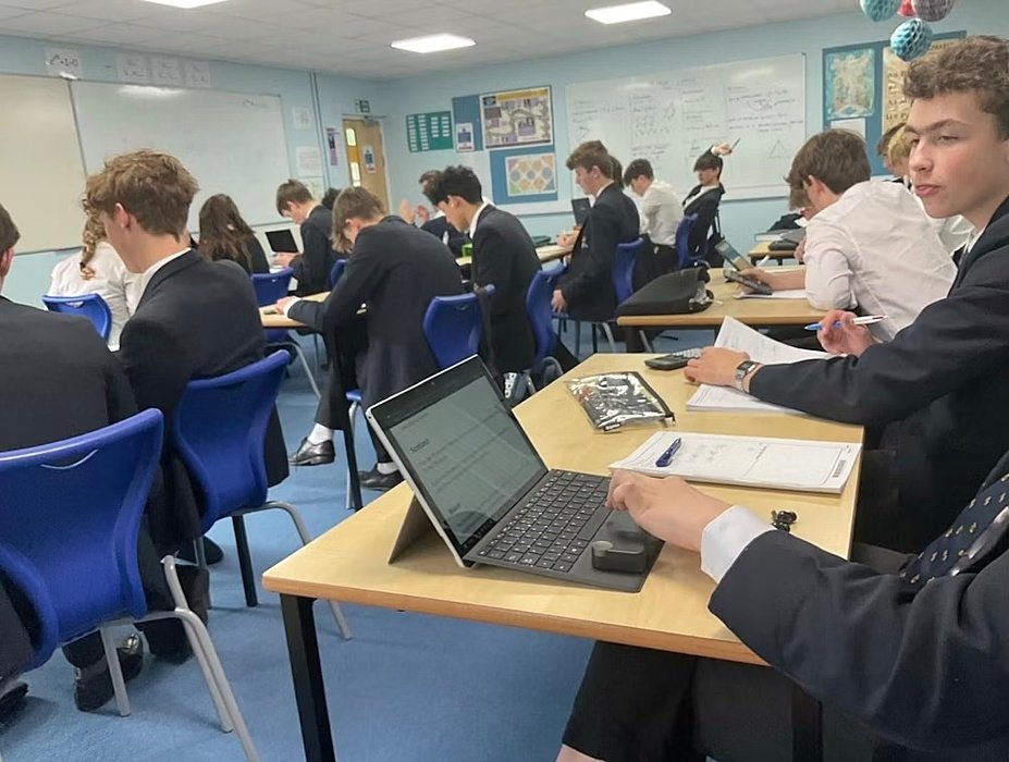 Classroom filled with students in uniform working on laptops and notebooks during a lesson.