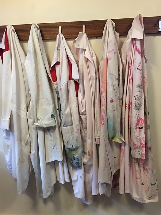 Row of paint-splattered white lab coats hanging on hooks along a wooden rack.