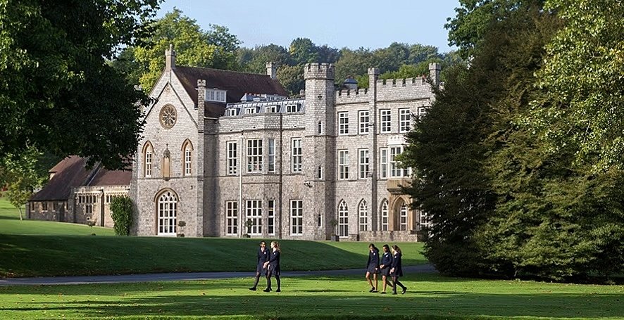 Large historic school building surrounded by trees with students walking across the green lawn in front.