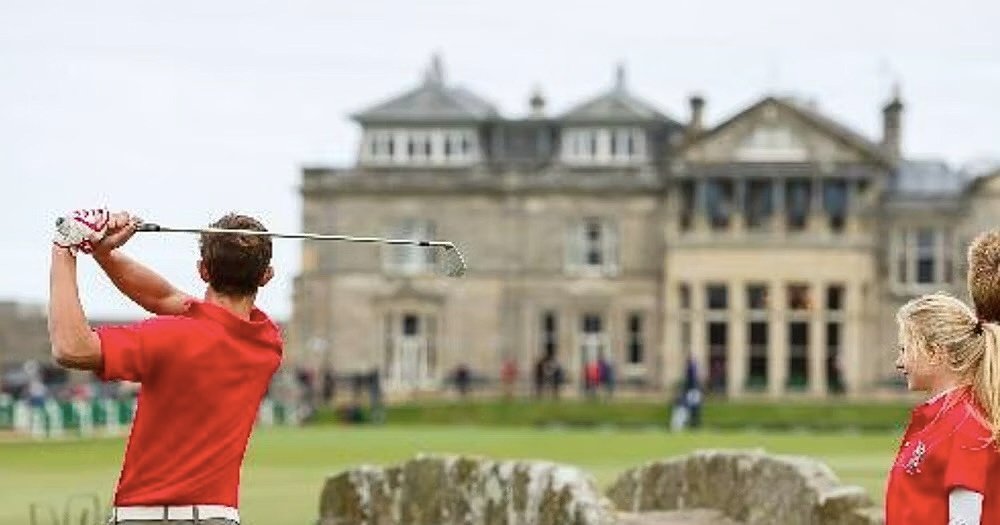 A male and a female golf player in red shirts with their clubs in the front and their school building in the background