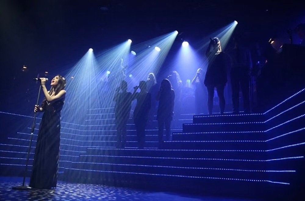 A singer performing under bright stage lights with a group of people standing in the background on illuminated steps.