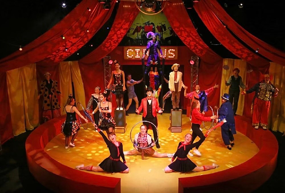 A group of performers in a circus-themed stage production with vibrant red curtains and spotlights.
