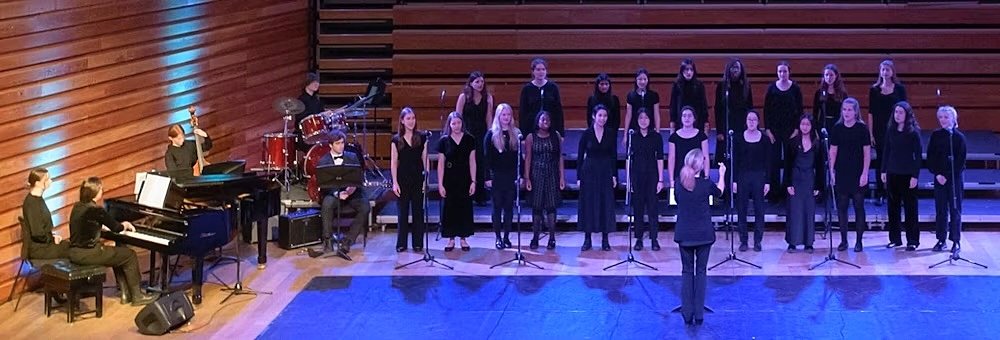 A choir of students performing on stage accompanied by a pianist and a drummer.