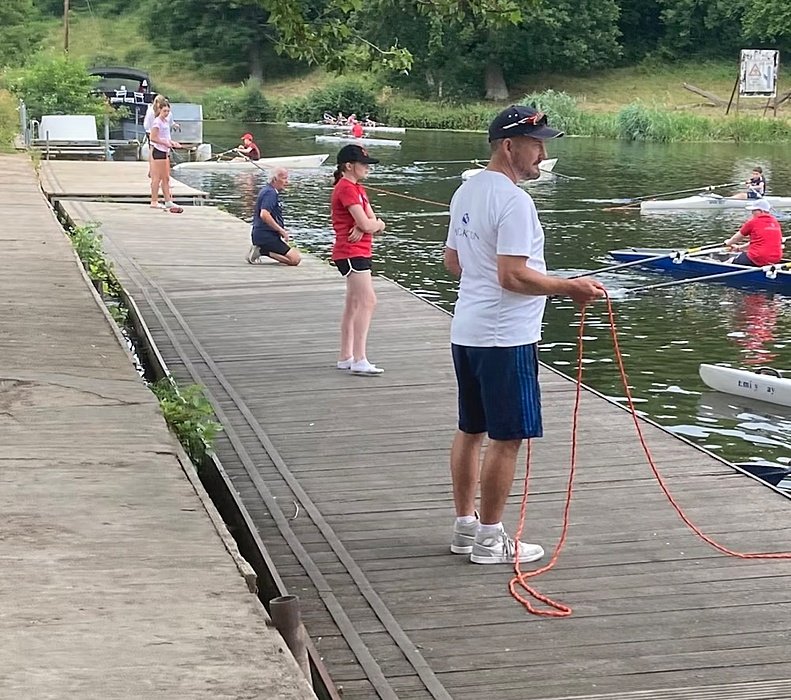 Students and a teacher on a landing in front of them rowing boats in the water