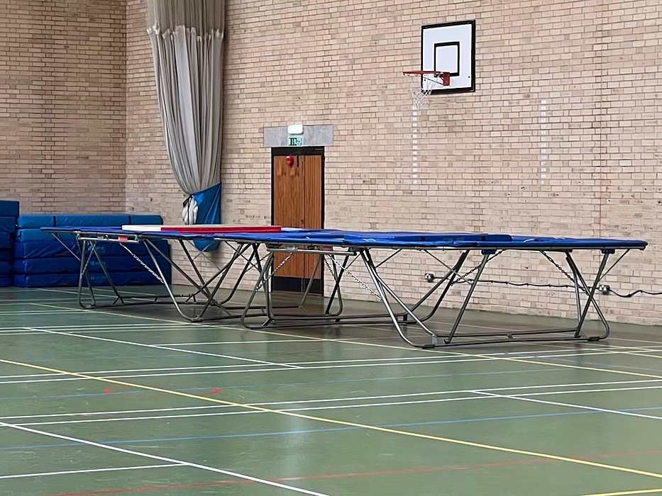 Indoor sports hall with trampolines, gym mats, and a basketball hoop on a brick wall.