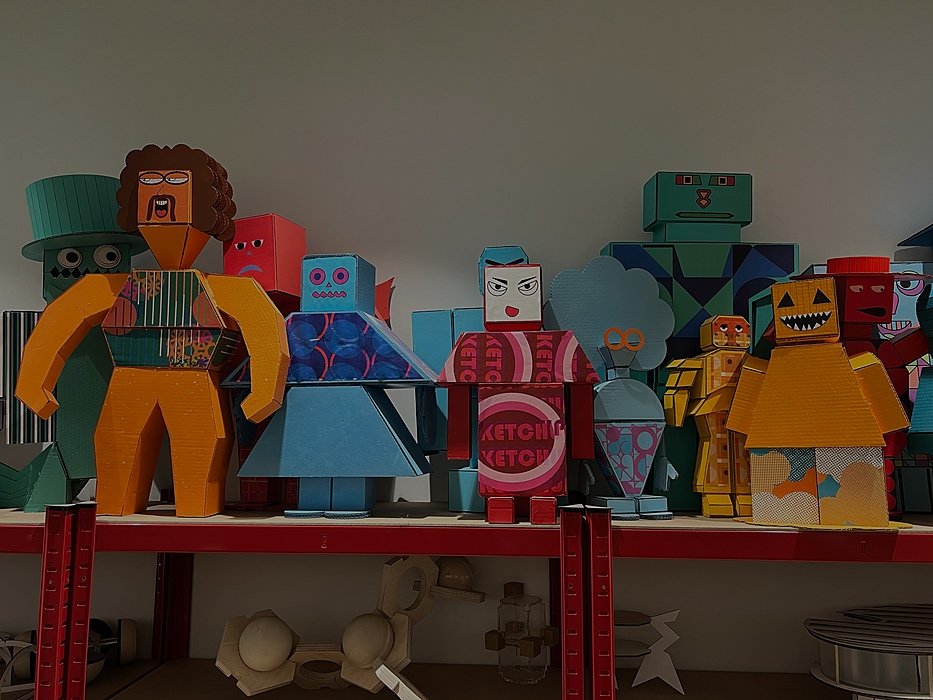 Colorful cardboard sculptures of stylized human and robot figures displayed on red shelves in an art classroom.