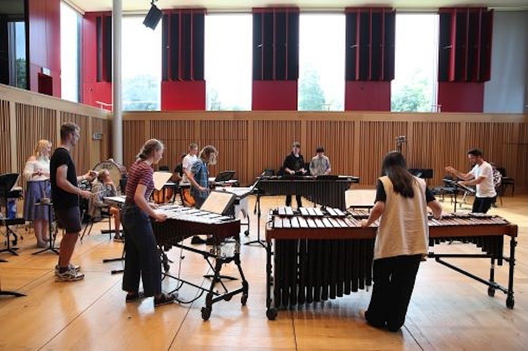 A group of students rehearsing on marimbas and other percussion instruments in a spacious, wood-paneled music room with large windows.