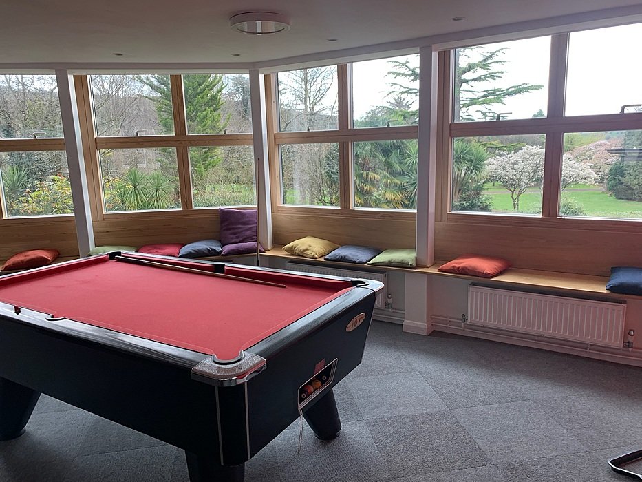A red pool table stands in front of large windows with a cushioned bench and colourful pillows.