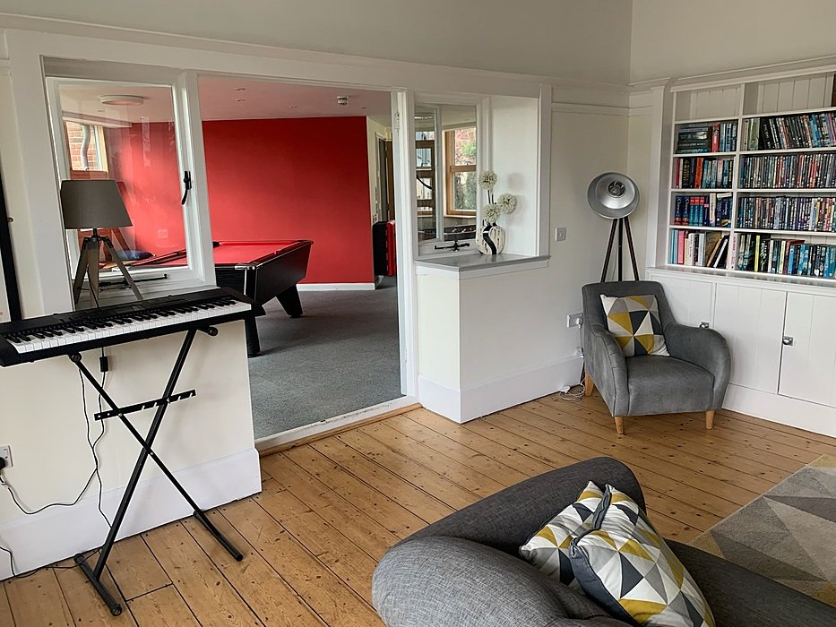 A living area with wooden flooring, grey armchairs and a doorway leading to a red room with a pool table.