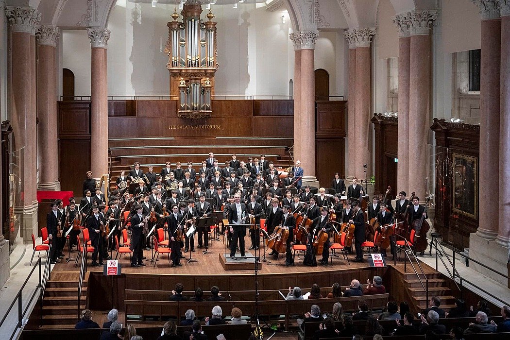 Large student orchestra performing in a grand hall with an organ and seated audience.
