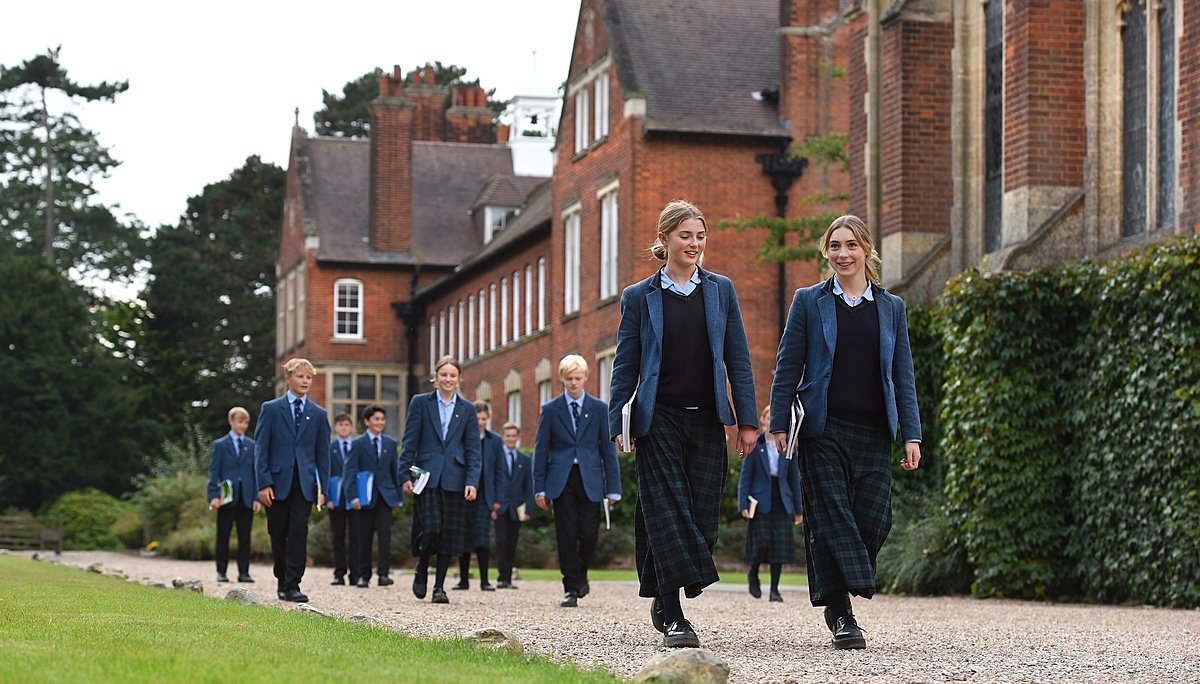 Students in blue blazers walking together on a school campus with historic red-brick buildings.