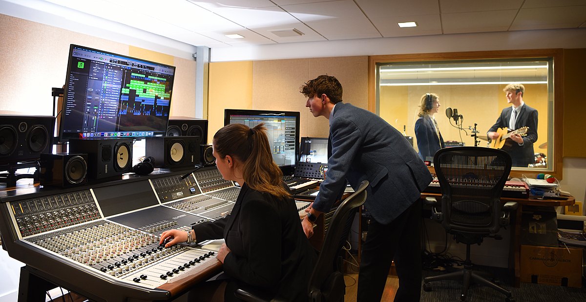 Students working in a music recording studio with sound equipment and a singer with a guitar in a booth.