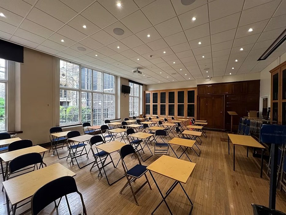 Bright classroom with wooden floors, large windows, and rows of exam desks.