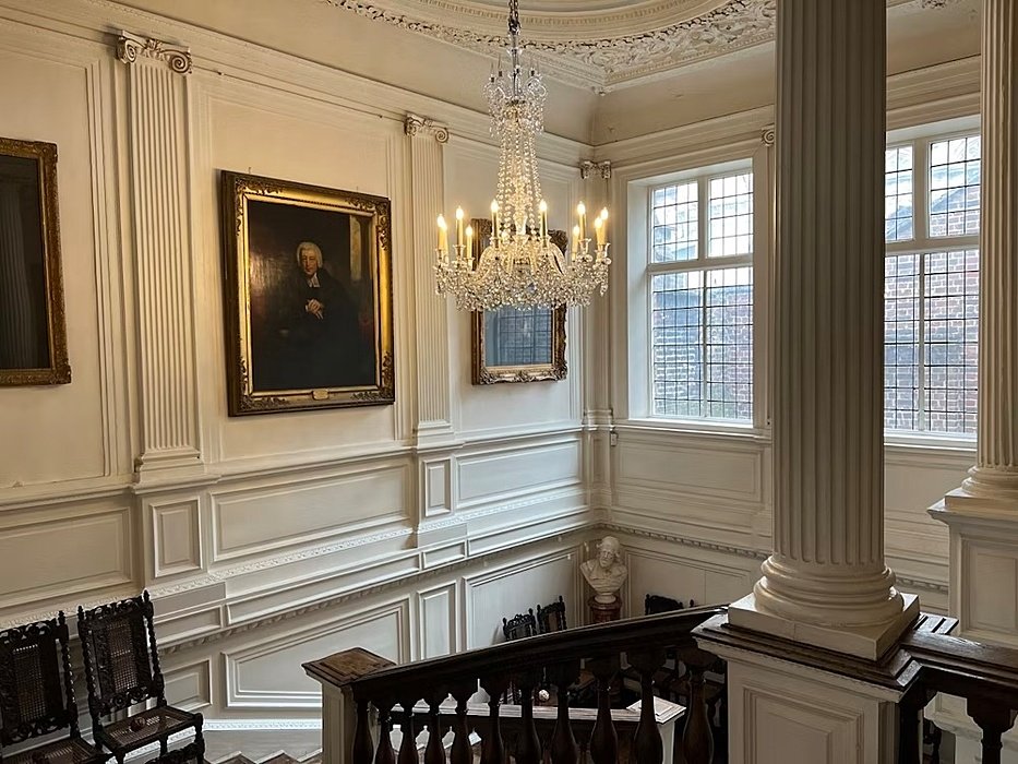 Elegant staircase with a chandelier, framed portraits, and tall windows letting in natural light.
