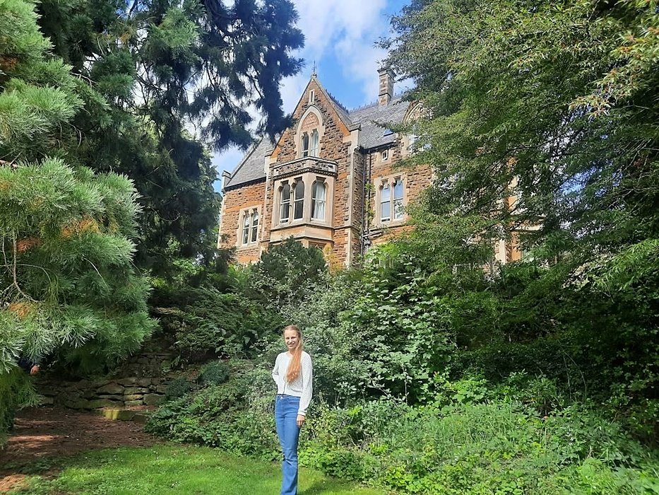 A blond girl with long hair in a jeans and jumper standing in a park in front of an old school building