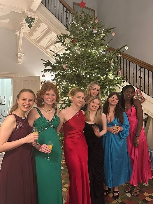 Seven girls in festive dresses smiling into the camera with a Christmas tree in the back