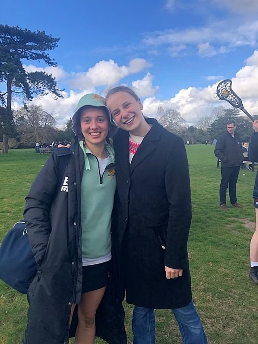 two girls arm in arm outside at a lacrosse match smiling into the camera