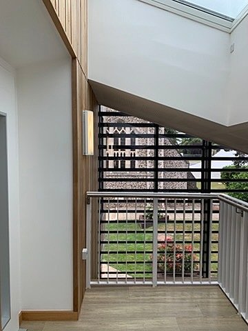 Modern staircase corner with wooden wall panels, metal railing, and a large window overlooking a garden and stone building.