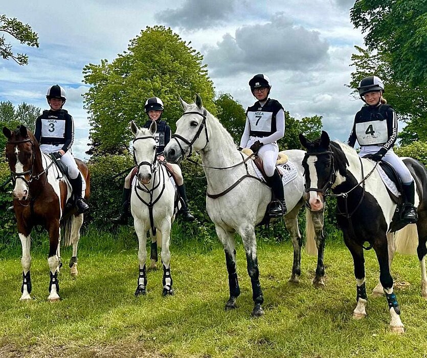 Four riders in equestrian gear sitting on their horses in a grassy field on a cloudy day.